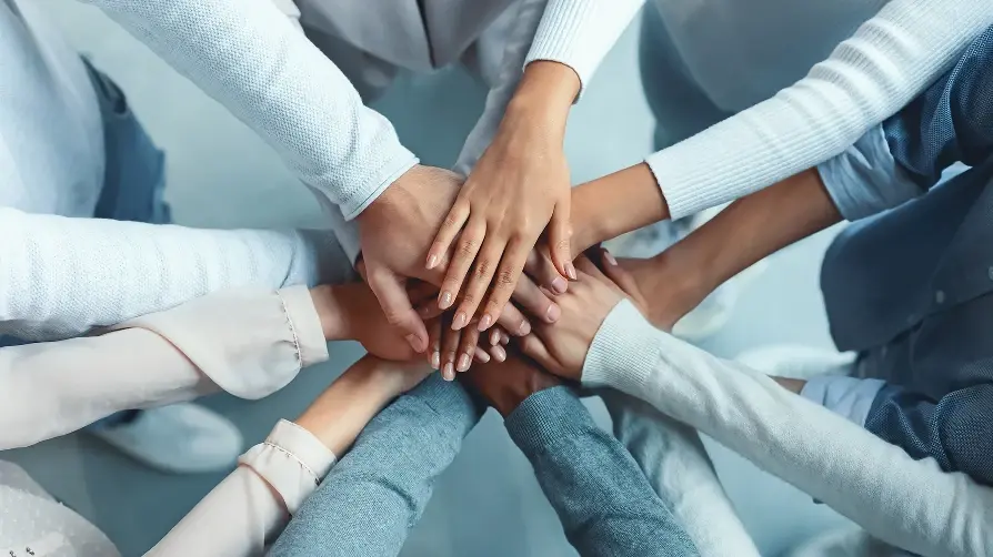 A group of people wearing light-colored sweaters and shirts, joining their hands together in a central stack.