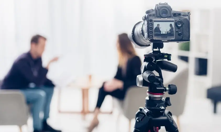 A an out-of-focus image of a woman sitting on a chair, with a camera stand in the foreground.