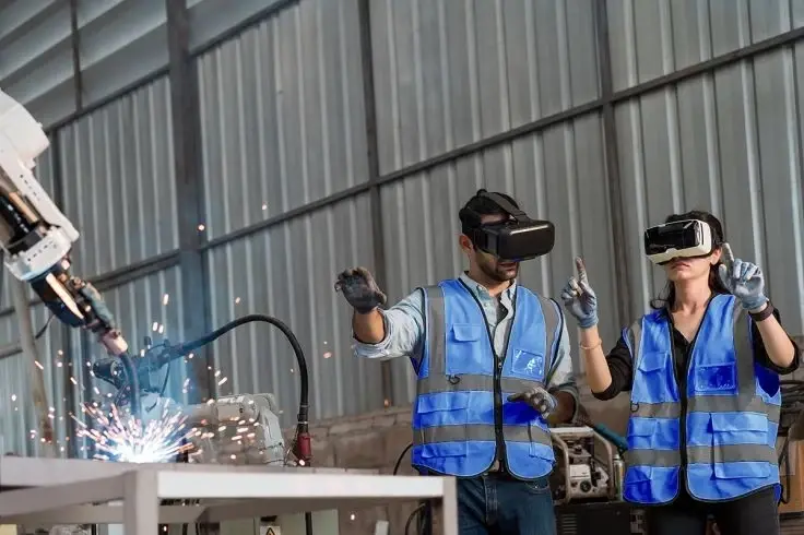 Two workers wearing VR headsets and safety vests monitor a robotic arm welding metal with sparks in a factory setting.