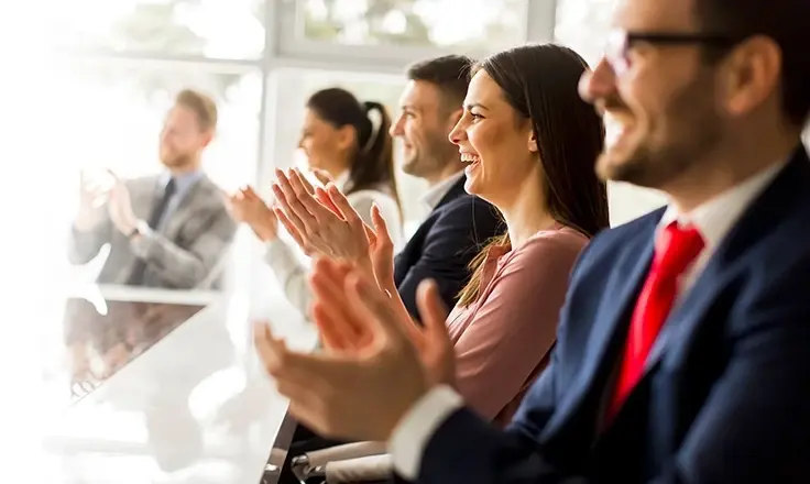 A group of smiling business professionals sitting in a row and clapping during a presentation in a bright office environment.