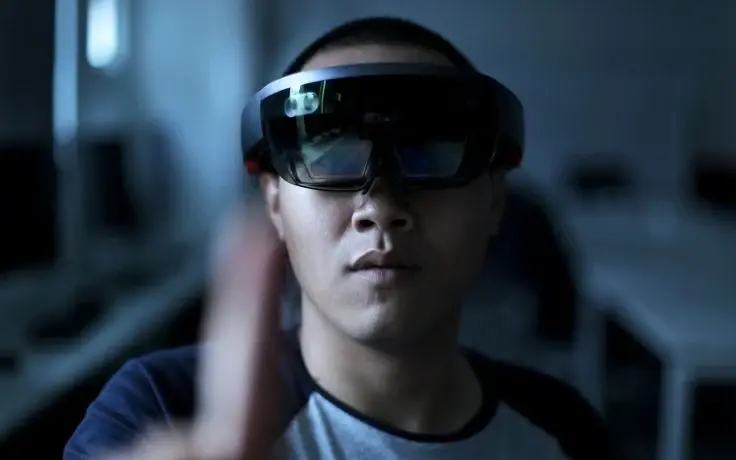A close-up of a person wearing a mixed reality headset while pointing a finger toward the camera in a dark room.