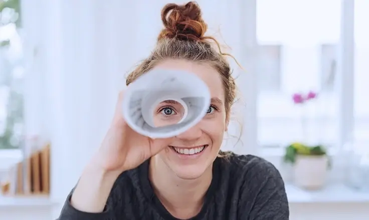 A smiling woman looks through a rolled-up paper tube like a telescope, with her blue eye visible in the center.