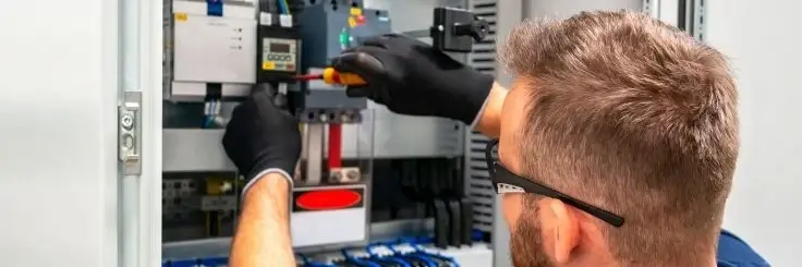 A technician in black gloves and safety glasses fixing an industrial electrical panel.