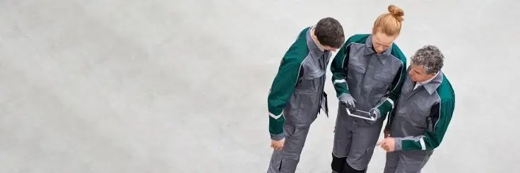 High-angle view of three workers in grey and green uniforms looking at a digital tablet together on a concrete floor.