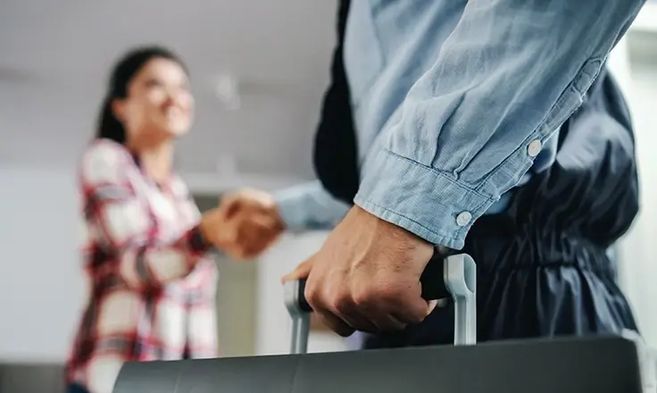 Close-up of a hand holding a tool case handle with a blurred woman and person shaking hands in the background.