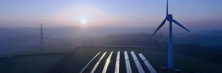 A large wind turbine and rows of solar panels in a field at sunrise with an electric pylon in the background.