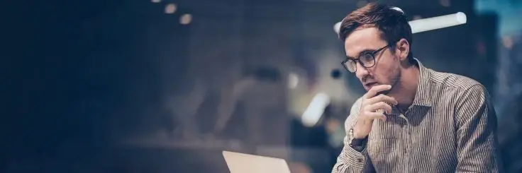 A man with glasses in a striped shirt looks thoughtfully at his laptop screen against a blurred office background.