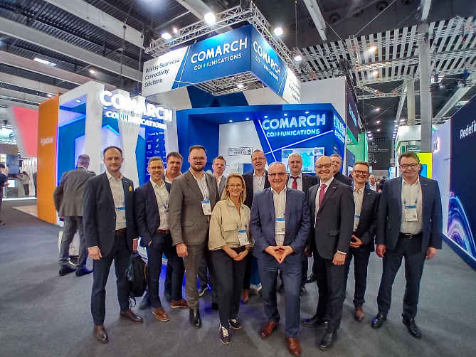 A large group of people in business attire pose together in front of a blue Comarch Communications trade show booth.