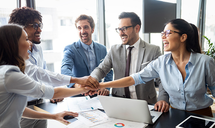 A multi-ethnic business team of five people smiling, laughing, and huddling with their hands stacked over a desk.